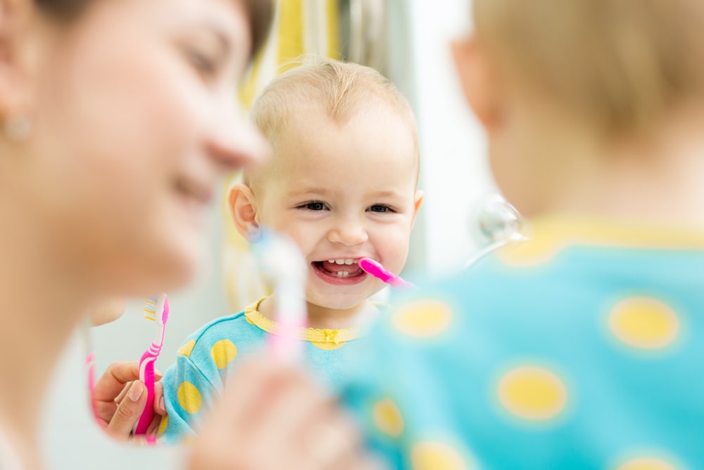 bambino in età pediatrica si lava i denti da latte con lo spazzolino davanti allo specchio insieme alla mamma